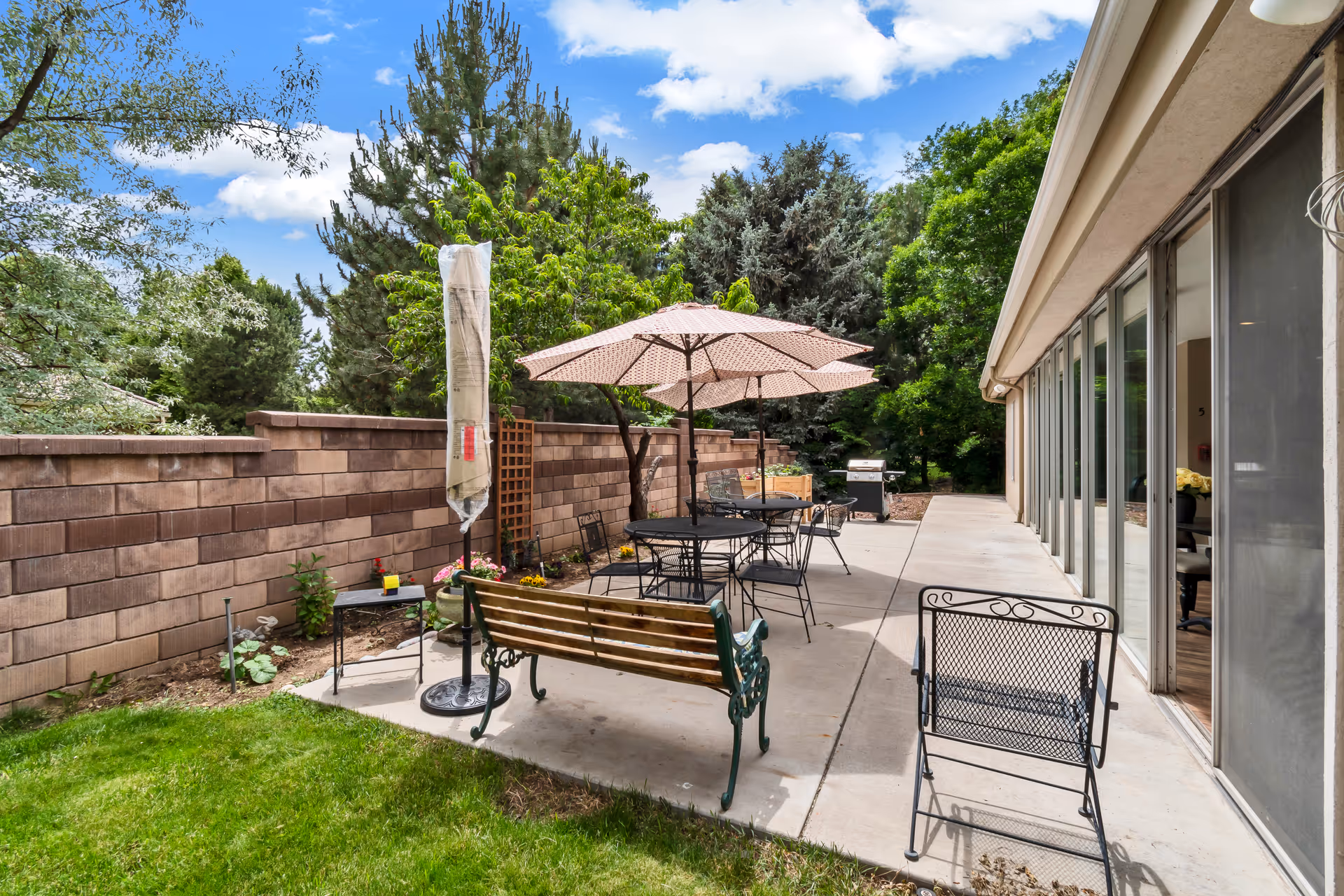 Outdoor patio area at Gardens Care Senior Living - Jewell Estates featuring a concrete walkway with metal tables and chairs, a wooden bench, a closed umbrella, and a barbecue grill. The area is surrounded by a brick wall and lush green trees under a partly cloudy sky.