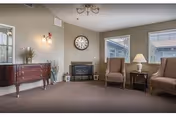 A cozy living room with two upholstered armchairs, a small wooden side table with a lamp, a wall clock above a fireplace, a wooden dresser with decorative items, and two windows letting in natural light.