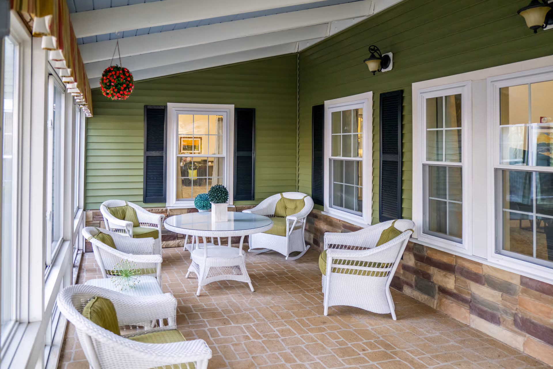 An enclosed porch with white wicker chairs and a round glass table against green siding and multiple windows.