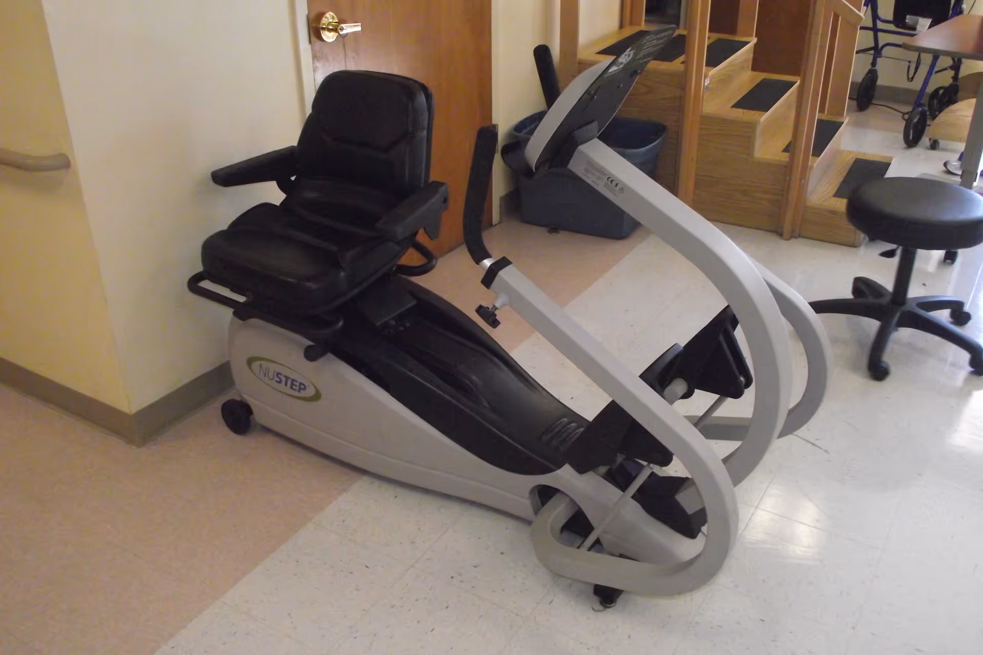 A NuStep recumbent cross trainer exercise machine with a black cushioned seat and armrests, placed indoors on a tiled floor near a wooden door and a mirror.
