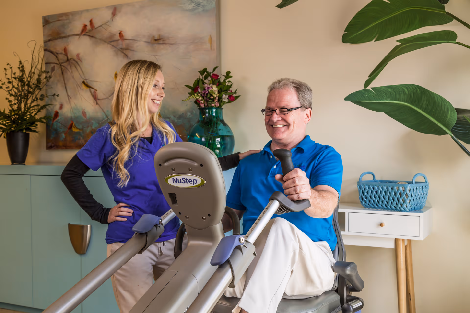A smiling elderly man using a NuStep exercise machine while a female caregiver or therapist stands beside him, offering support and encouragement in a well-decorated room with plants and artwork.