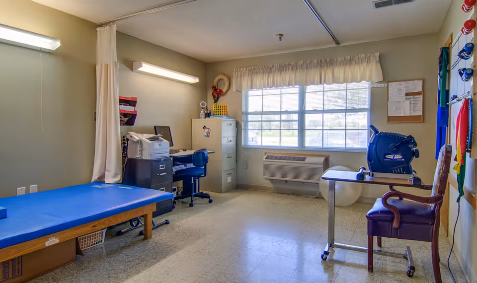 A therapy or rehabilitation room with a blue padded treatment table on the left, a desk with a computer and office chair, a filing cabinet, and a window with blinds and a valance. On the right side, there is a small table with exercise equipment and a chair. The walls have various therapy tools and a bulletin board.