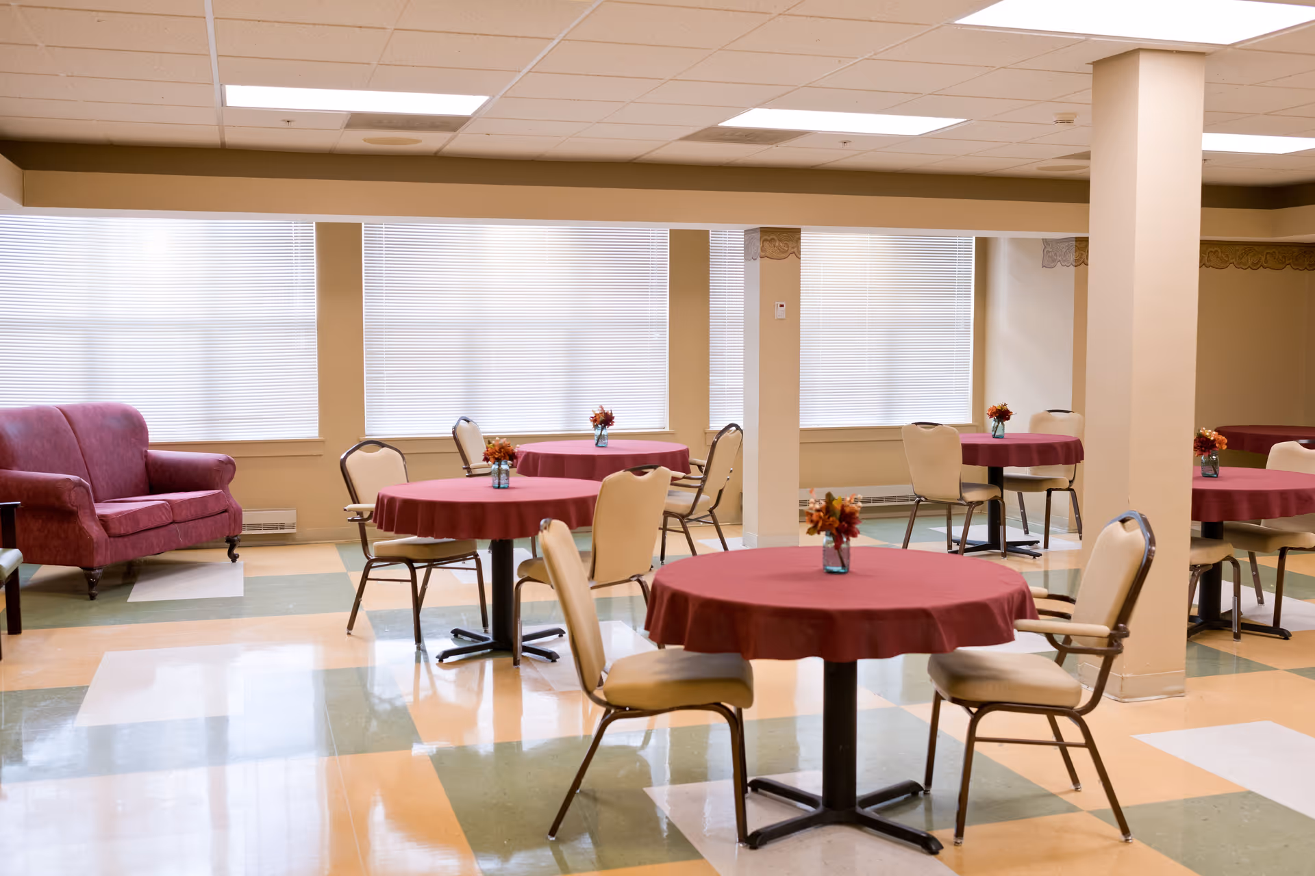 A dining area with several round tables covered with maroon tablecloths, each table decorated with a small vase of flowers. Beige cushioned chairs surround the tables. There is a maroon loveseat against the wall with large windows covered by blinds. The floor has a checkered pattern with green, beige, and white tiles.