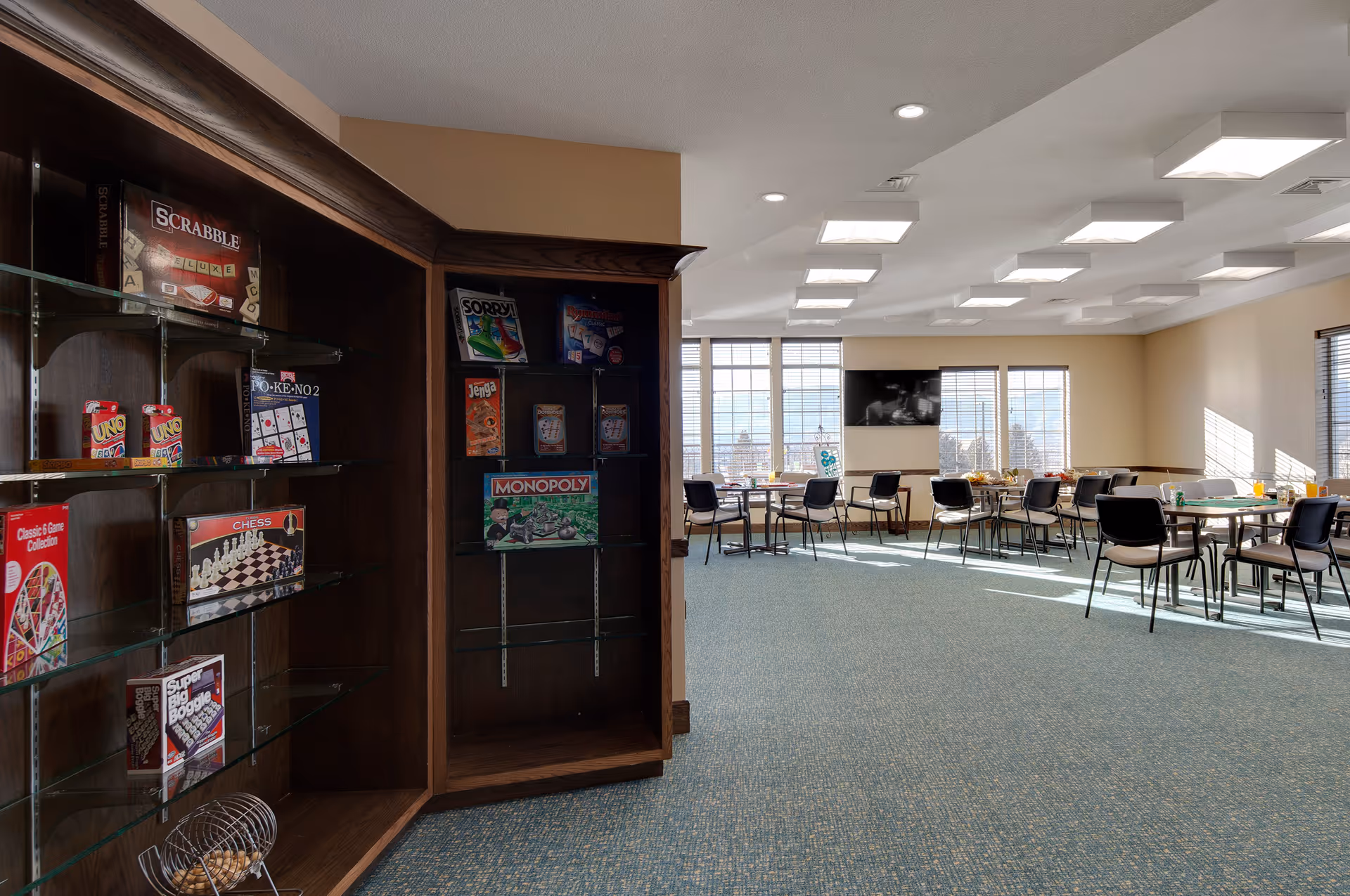 A bright and spacious activity room in a senior living facility with large windows letting in natural light. The room has several tables and chairs arranged for group activities or dining. On the left side, there is a wooden display cabinet with glass shelves holding various board games and card games such as Scrabble, Sorry, Rummikub, Jenga, Monopoly, Uno, Chess, and others. The walls are light-colored, and the ceiling has modern square light fixtures.