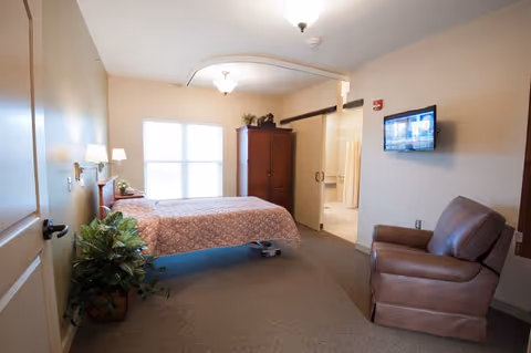 A well-lit bedroom in a senior living facility featuring a bed with a patterned bedspread, a large window with blinds, a wooden armoire, a brown leather armchair, a potted plant, and a wall-mounted television. The room has beige walls and carpeted flooring.