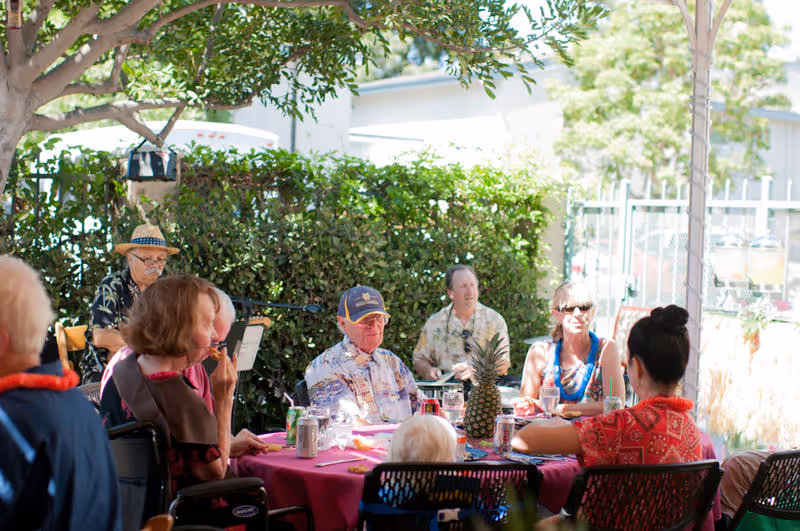 A group of elderly people and a caregiver sitting around a table outdoors under a canopy, enjoying a social gathering with drinks and a pineapple centerpiece. The setting is shaded by trees and surrounded by greenery.