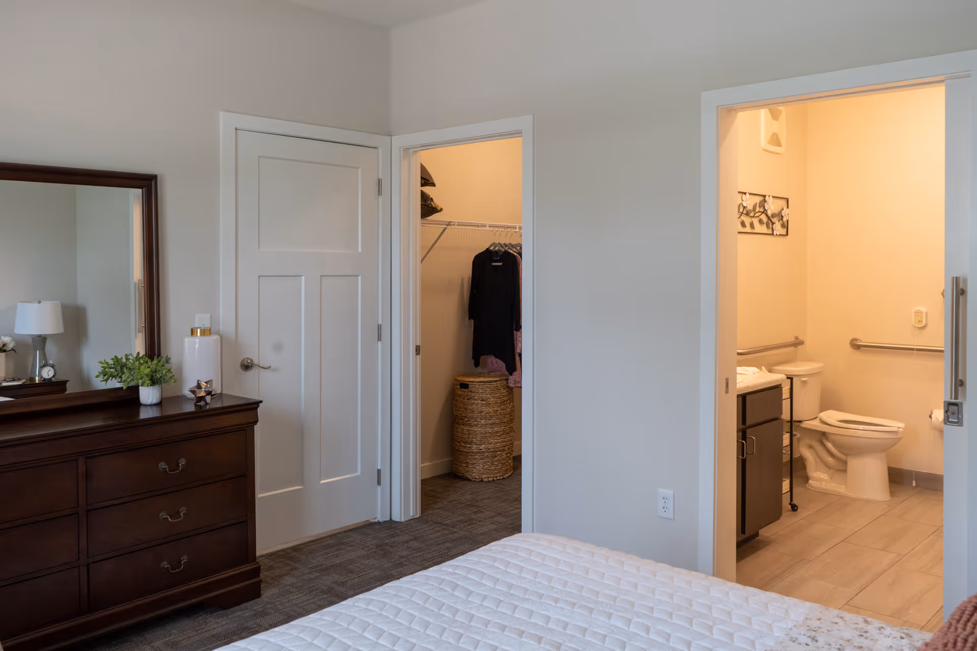 View of a bedroom in The Canopy at Azalea Grove showing a bed with a white quilted cover, a dark wooden dresser with a mirror, a small plant, and decorative items. There are two open doors: one leading to a walk-in closet with clothes hanging and a woven basket, and the other leading to a bathroom with a toilet, grab bars, and a vanity with a sink.