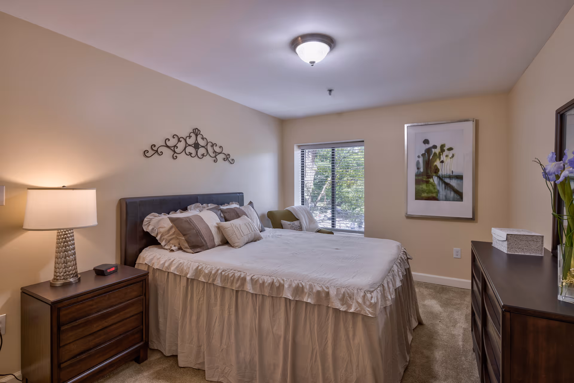 A comfortable bedroom with a made bed, bedside table and lamp, dresser, window, and framed artwork.