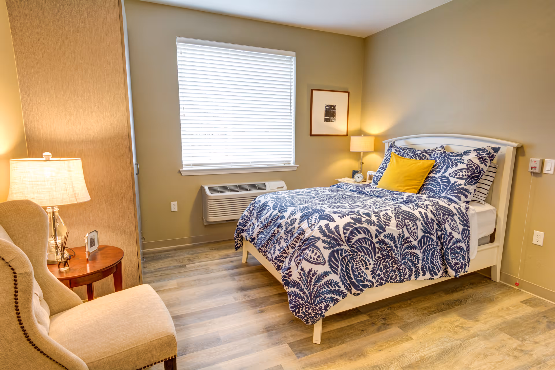 A cozy bedroom with a bed featuring blue and white patterned bedding and a yellow accent pillow. There is a beige upholstered chair next to a small round wooden side table with a lamp and a small framed photo. A window with closed blinds is on the wall behind the bed, and a framed picture hangs on the wall beside a second lamp on a nightstand.