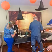 Three people standing around a table decorated with Halloween-themed items including witch hats hanging above and a backdrop with a spooky tree and orange sky. One person is holding a walker.