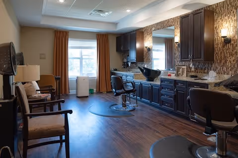Interior view of a salon area in a senior living facility with wooden flooring, dark wood cabinetry, a large mirror, salon chairs, and a window with orange curtains letting in natural light.