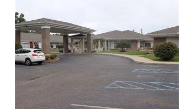 Exterior view of Holzer Senior Care Center showing a driveway with a covered entrance supported by columns, a white car parked on the left, and single-story buildings with a lawn and bushes under an overcast sky.