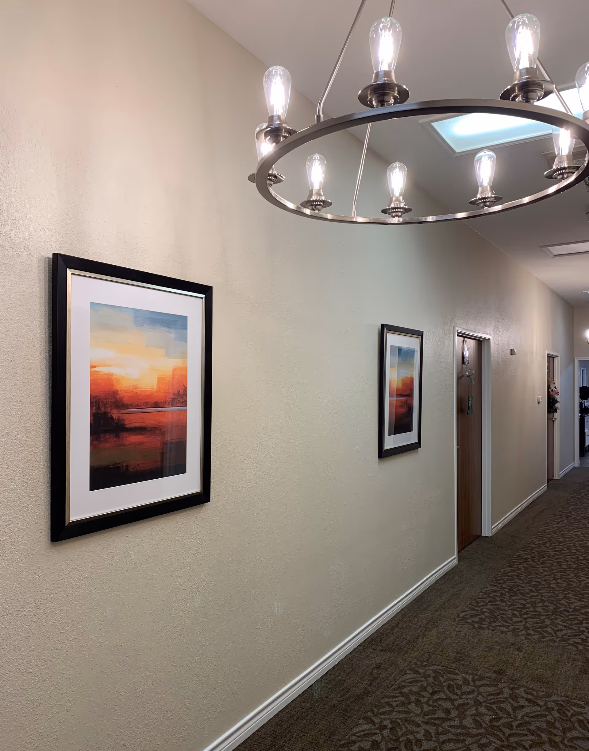 A hallway with beige walls and patterned carpet. Two framed abstract paintings with warm colors hang on the left wall. There are several wooden doors on the right side of the hallway. A modern circular light fixture with exposed bulbs hangs from the ceiling.