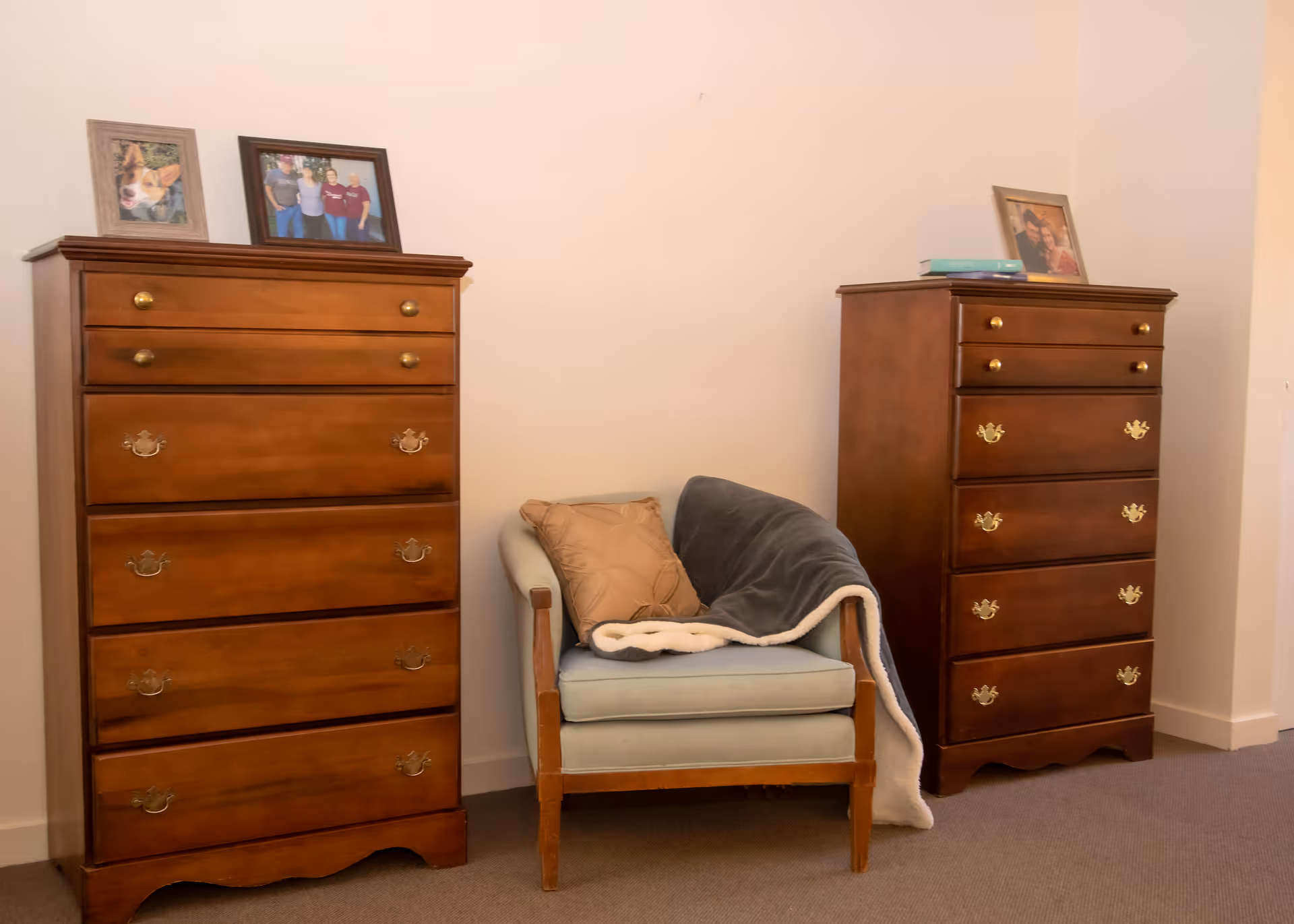 A cozy corner of a room featuring two wooden dressers with brass handles, each topped with framed photographs. Between the dressers is a light blue cushioned armchair with wooden arms, adorned with a beige pillow and a gray blanket draped over one side. The walls are plain and light-colored, and the floor is carpeted.