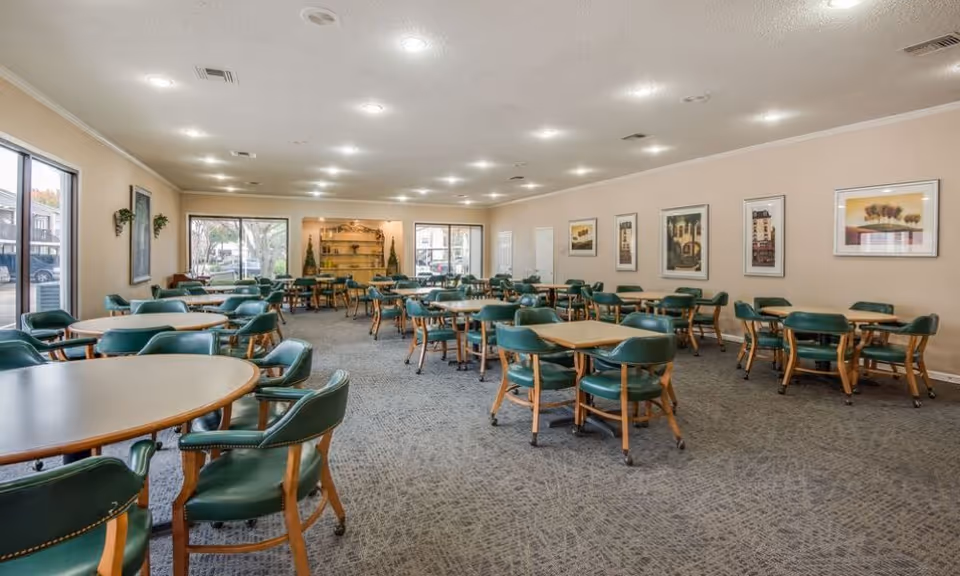 A spacious dining room with multiple round tables surrounded by green cushioned chairs with wooden legs. The room has beige walls adorned with framed artwork and large windows letting in natural light. The carpet is patterned in shades of gray, and the ceiling has recessed lighting.