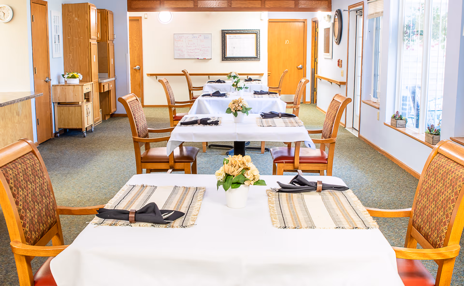 A bright dining room with several tables covered in white tablecloths, each set with placemats, black napkins, and small flower arrangements. Wooden chairs with patterned upholstery surround the tables. Large windows on the right side let in natural light, and wooden cabinets and doors are visible along the left and back walls.