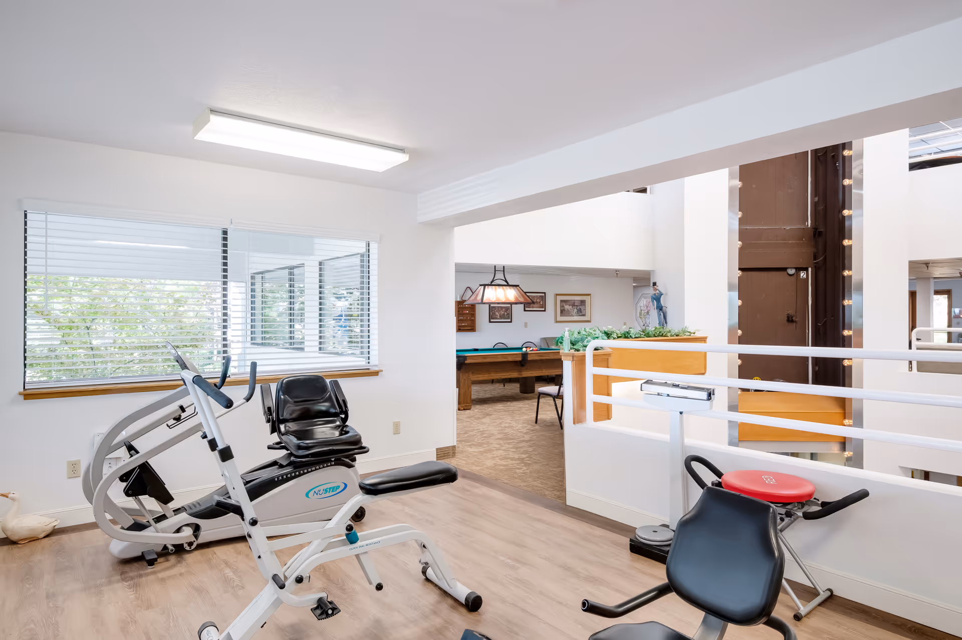 A bright exercise room with fitness equipment including a recumbent bike and a rowing machine. Large windows with blinds allow natural light to enter. In the background, there is a recreational area with a pool table and framed pictures on the wall.