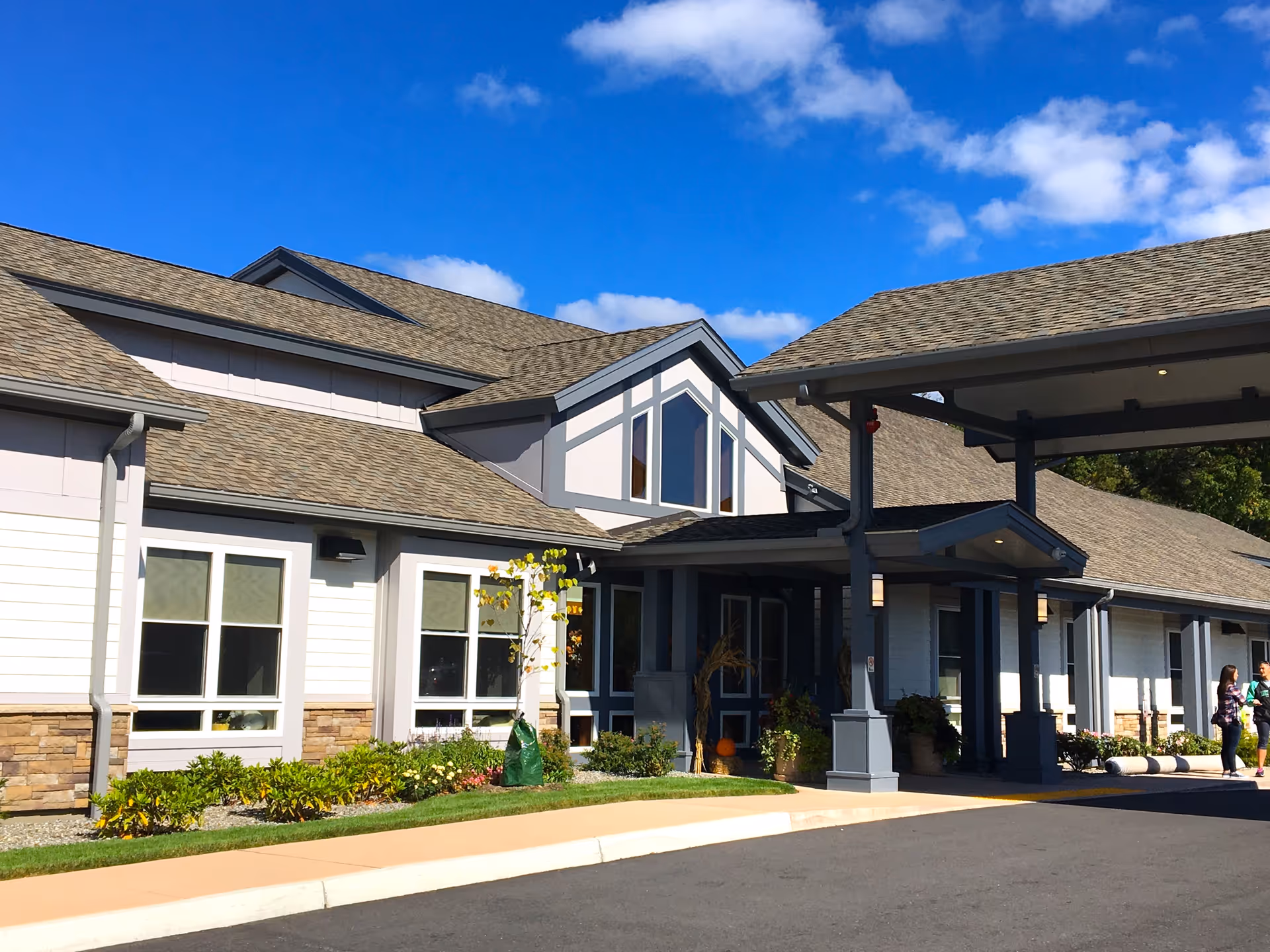 Front entrance of a residential care building with a covered drop-off, landscaped beds, and a bright blue sky.