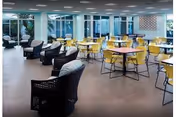 A spacious, modern common area with multiple round tables surrounded by yellow chairs and several black cushioned armchairs. Large windows allow natural light to fill the room, and there is a decorative wall piece visible in the background.