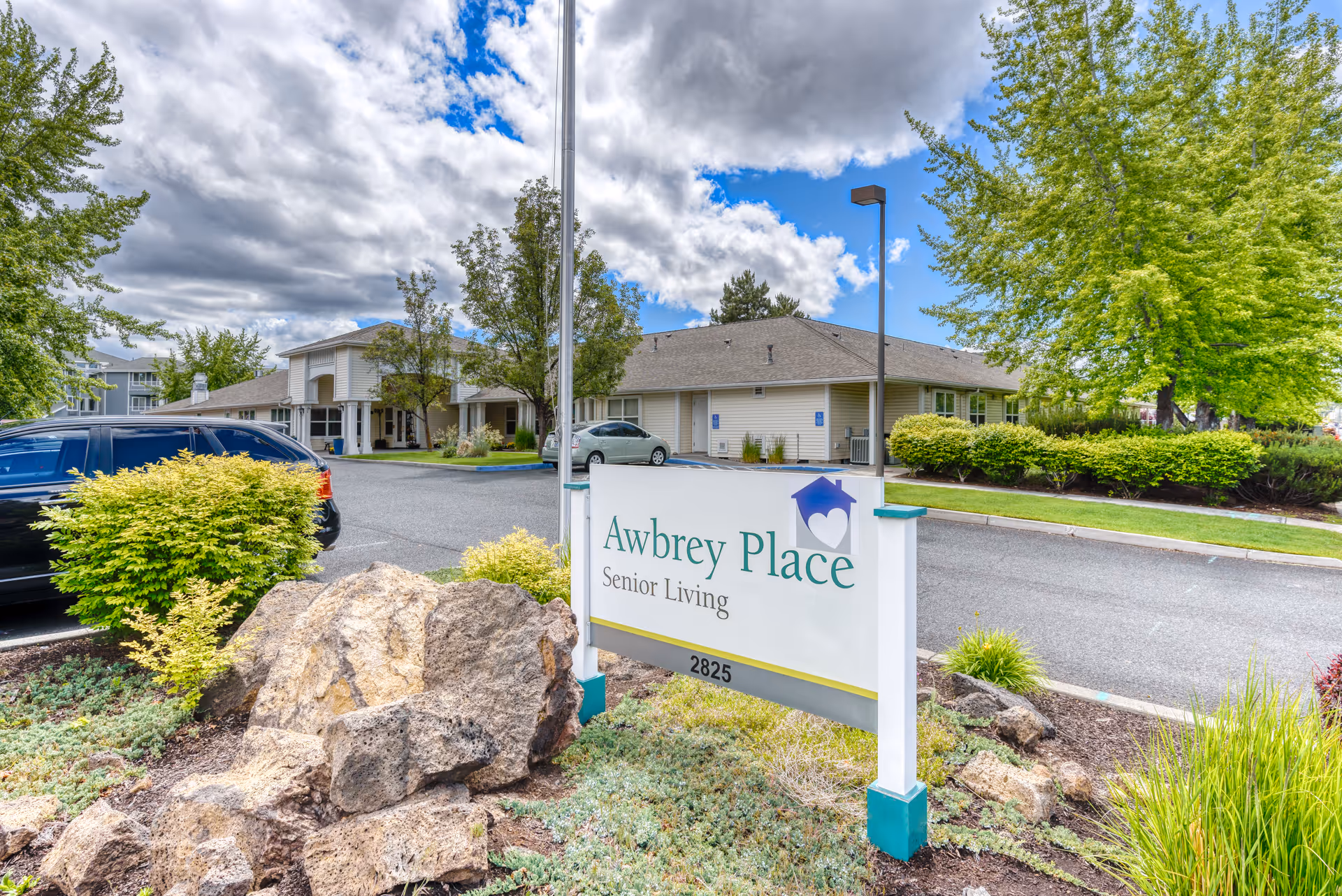 Outdoor view of Awbrey Place Senior Living facility showing the entrance sign with landscaping including rocks, bushes, and trees. The building and parking area are visible under a partly cloudy sky.