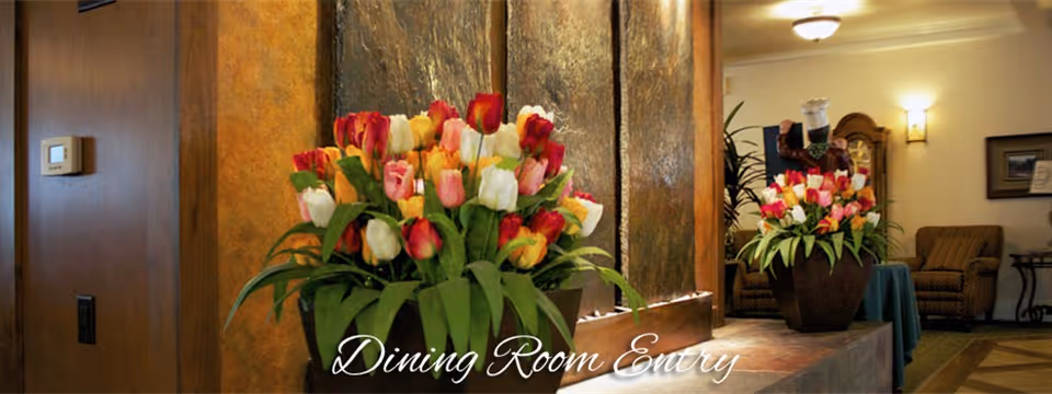 Interior view of a dining room entry area with a large flower arrangement of colorful tulips in the foreground, a water feature behind it, and seating with armchairs and a table in the background.