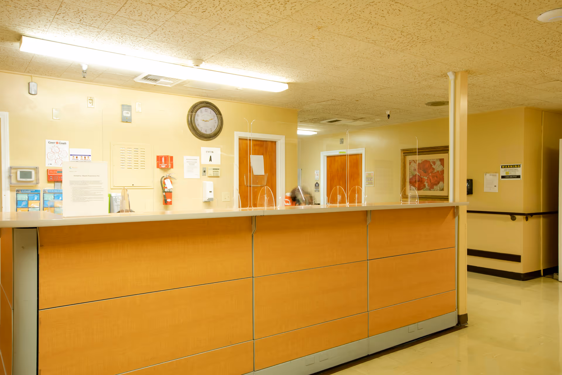 Reception desk area inside a senior living facility with a wooden front counter, clear protective barriers, a wall clock, fire extinguisher, informational posters, and doors leading to other rooms. The walls are painted light yellow and there is a framed floral picture on one wall.