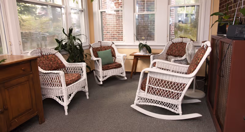 A cozy sitting area with four white wicker chairs featuring floral cushions arranged around a small wooden table with a potted plant. The space is enclosed by windows showing a brick exterior wall and greenery outside. There is a wooden cabinet on the left and a standing fan on the right.