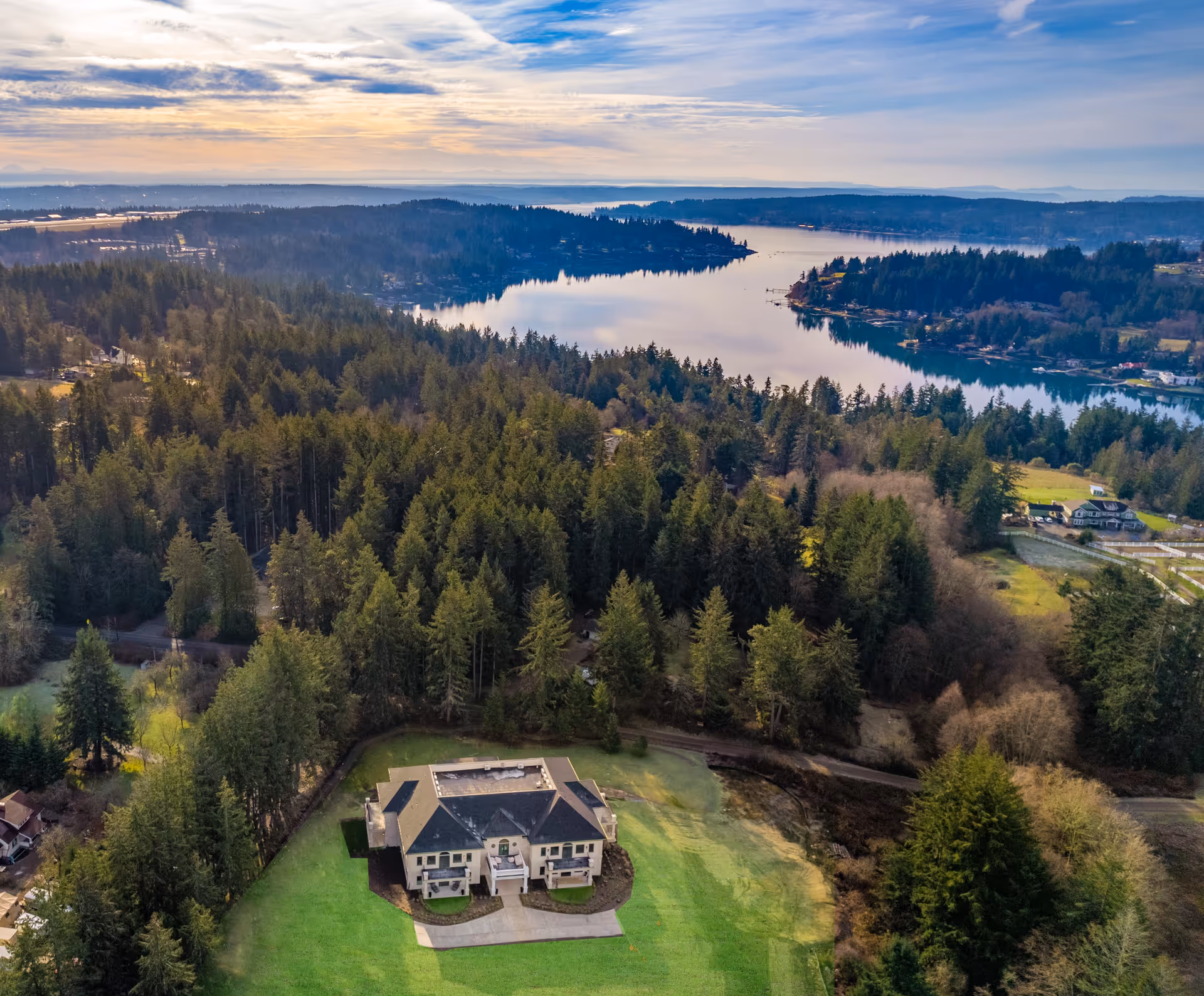 Aerial view of a large residential building on a grassy lawn surrounded by dense forest with a lake and hills in the background.