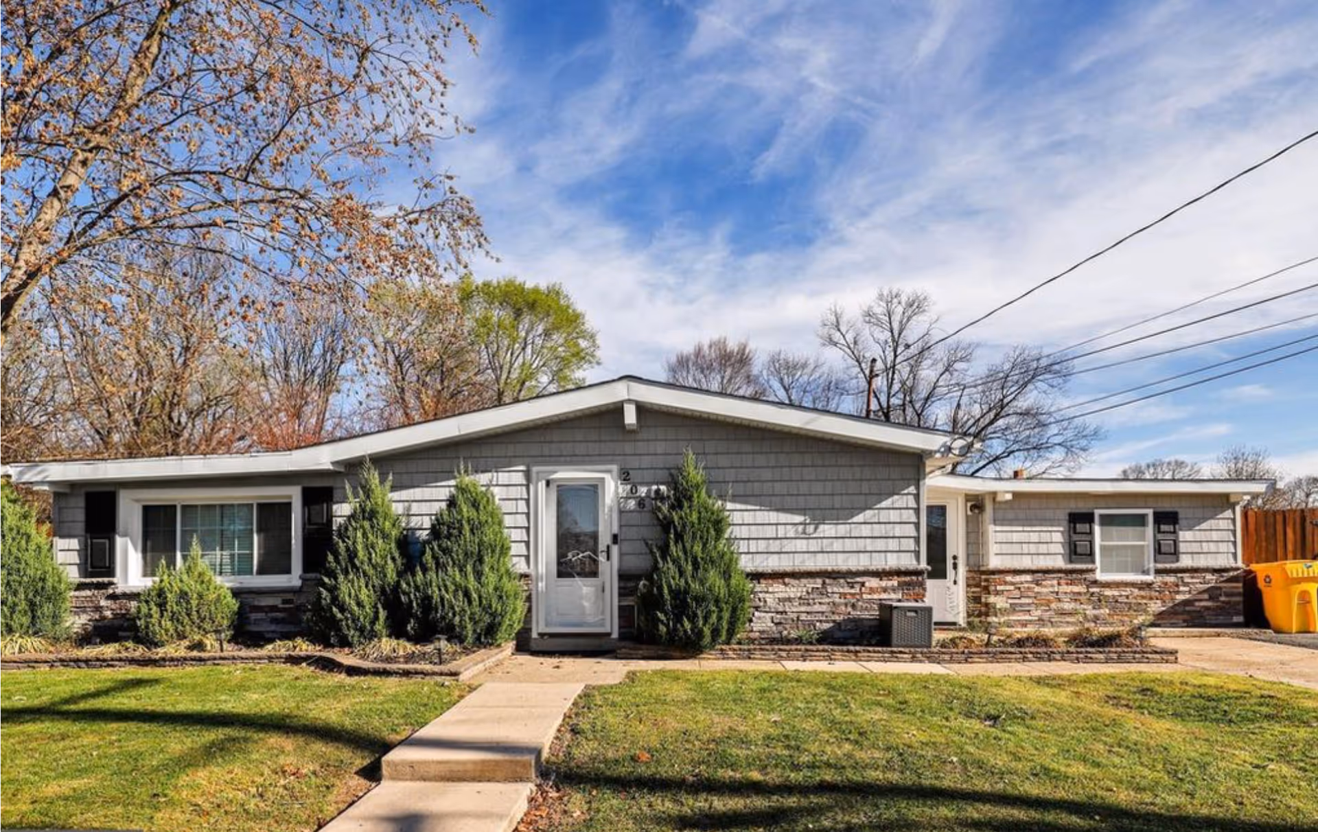 Single-story residential building with gray siding and stone accents, a white front door, and several windows with black shutters. There are green shrubs and a well-maintained lawn in front, with a concrete walkway leading to the entrance. Trees with sparse leaves and a blue sky with some clouds are visible in the background.