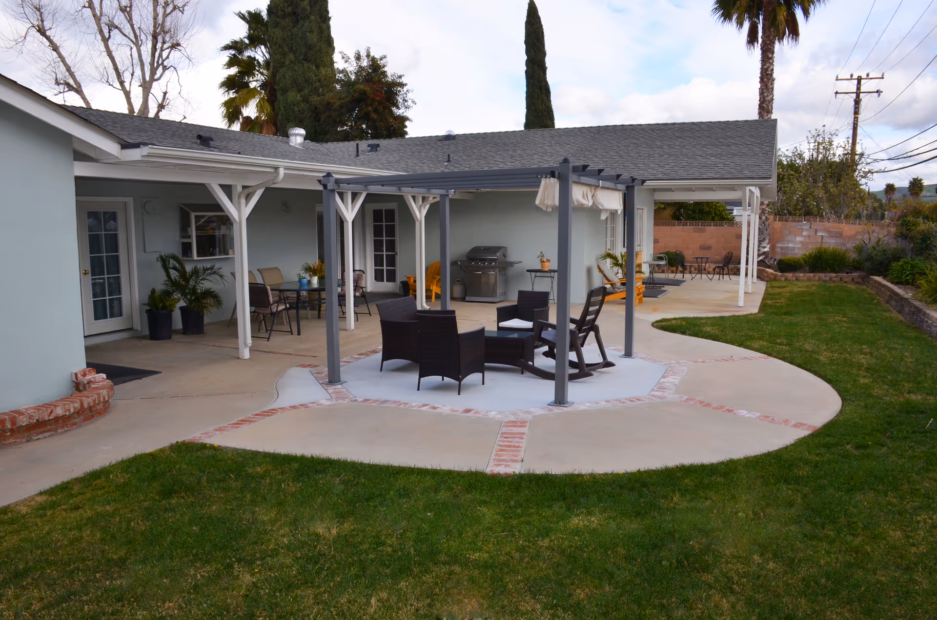 Outdoor patio area at a senior living facility with a covered seating area featuring wicker chairs and a small table, a barbecue grill, additional seating under a covered porch, and a well-maintained lawn with trees and shrubs in the background.
