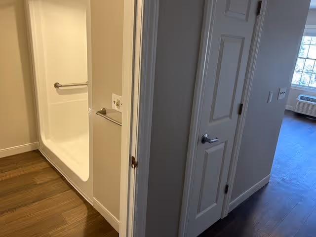 Interior view showing a hallway with a closed white door on the right and an open doorway on the left revealing a white bathtub with a grab bar. The floor is wooden, and a window with an air conditioning unit is visible in the background.