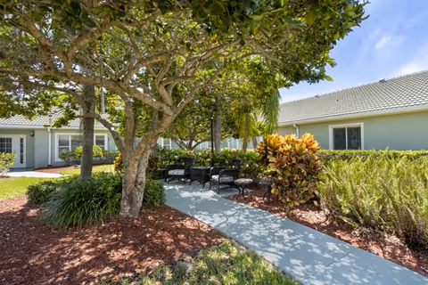 Outdoor seating area with black wicker chairs and a table under the shade of a large tree, surrounded by bushes and plants, with single-story buildings in the background under a clear blue sky.