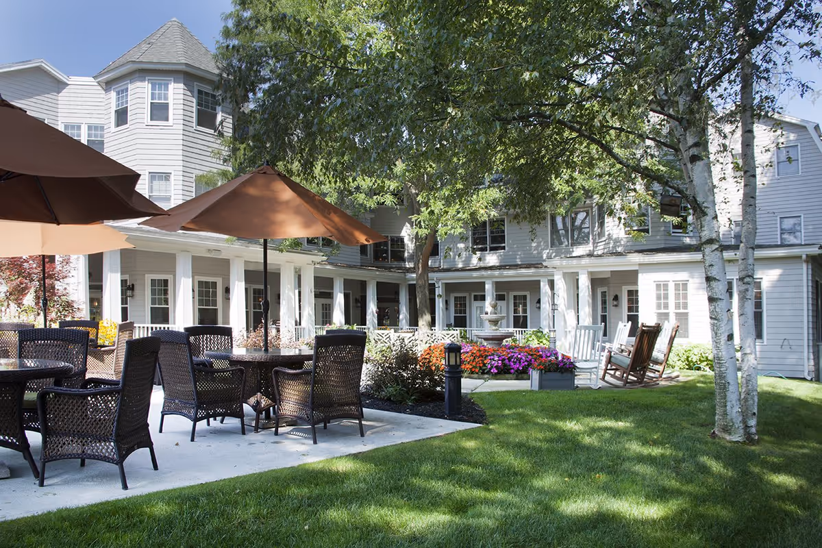 Courtyard patio with wicker tables and umbrellas, rocking chairs, a fountain and flowerbeds in front of a multi-story building.