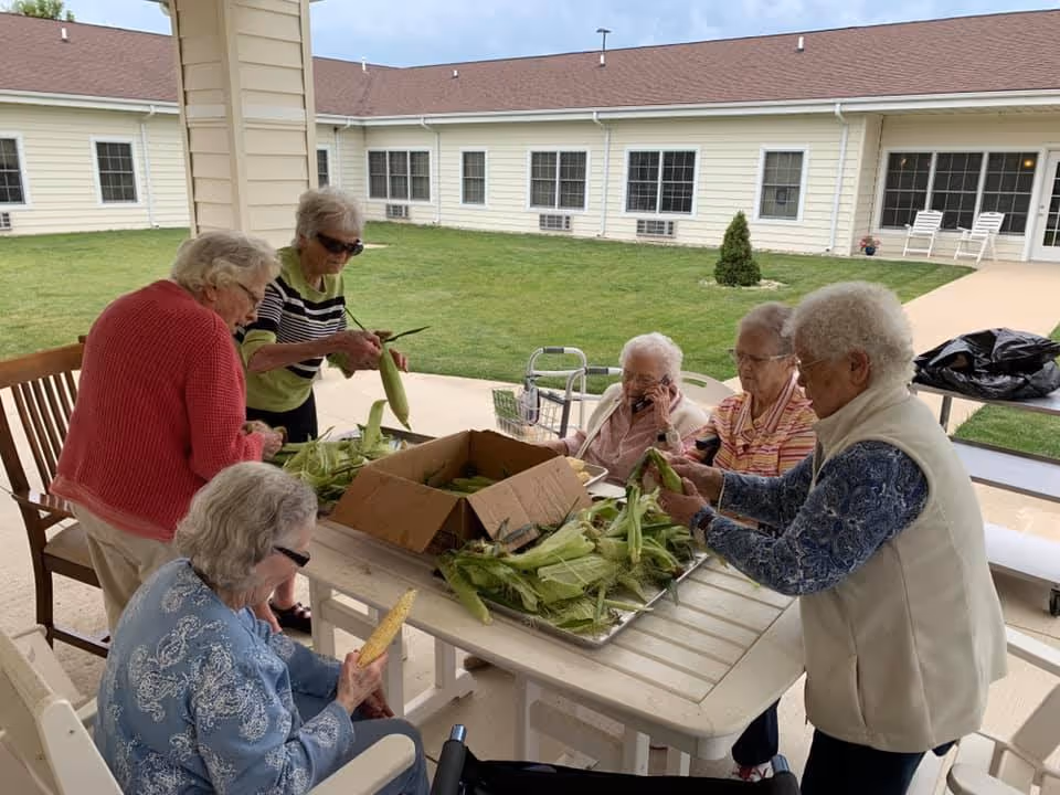 Five elderly women sitting and standing around a white outdoor table, husking fresh corn. They are on a covered patio area with a grassy courtyard and a building with multiple windows in the background.