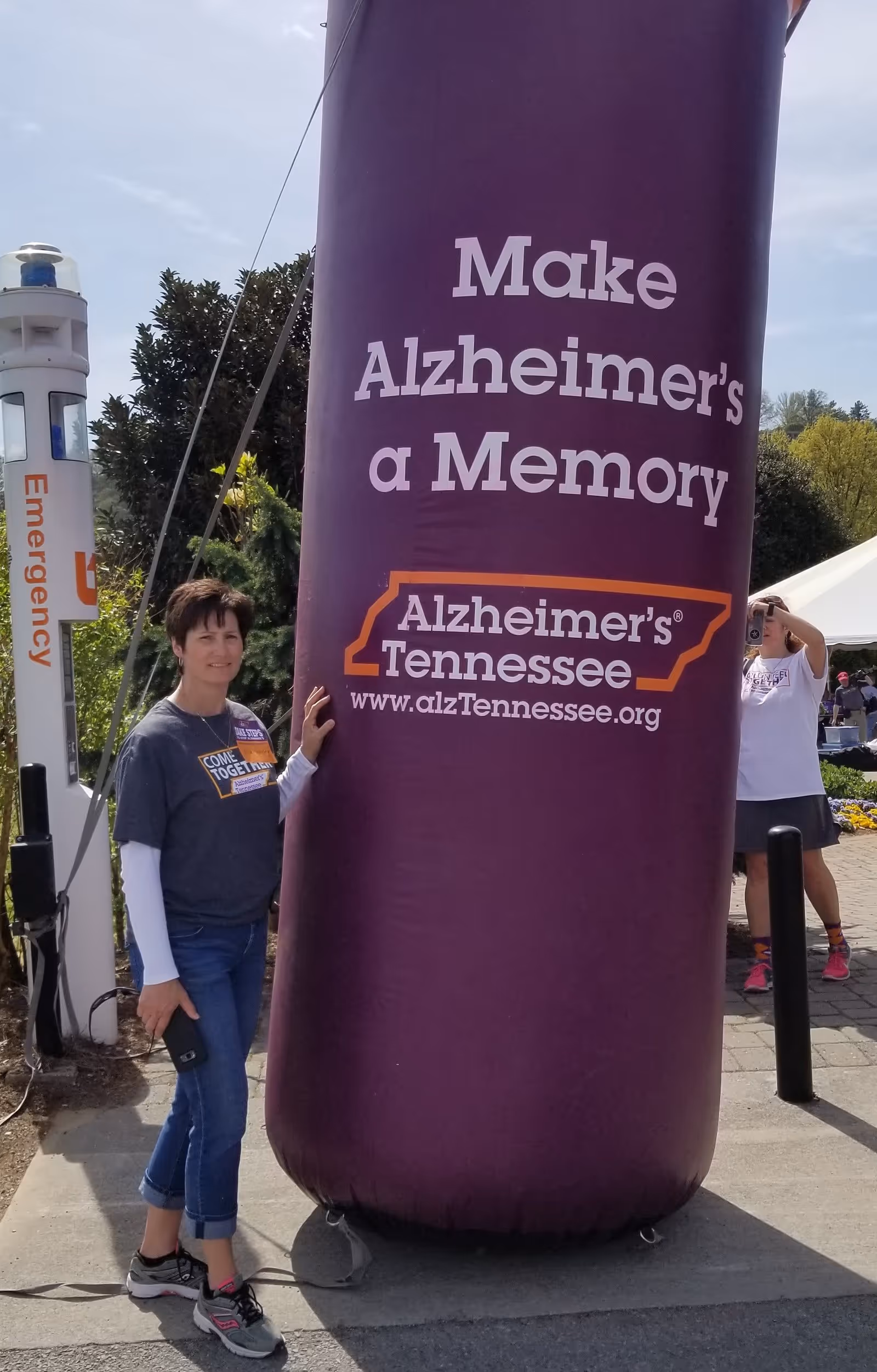 A woman standing outdoors next to a large purple inflatable pillar that reads 'Make Alzheimer's a Memory Alzheimer's Tennessee www.alzTennessee.org'. Another person in the background is taking a photo with a smartphone. There is an emergency call box visible on the left side.