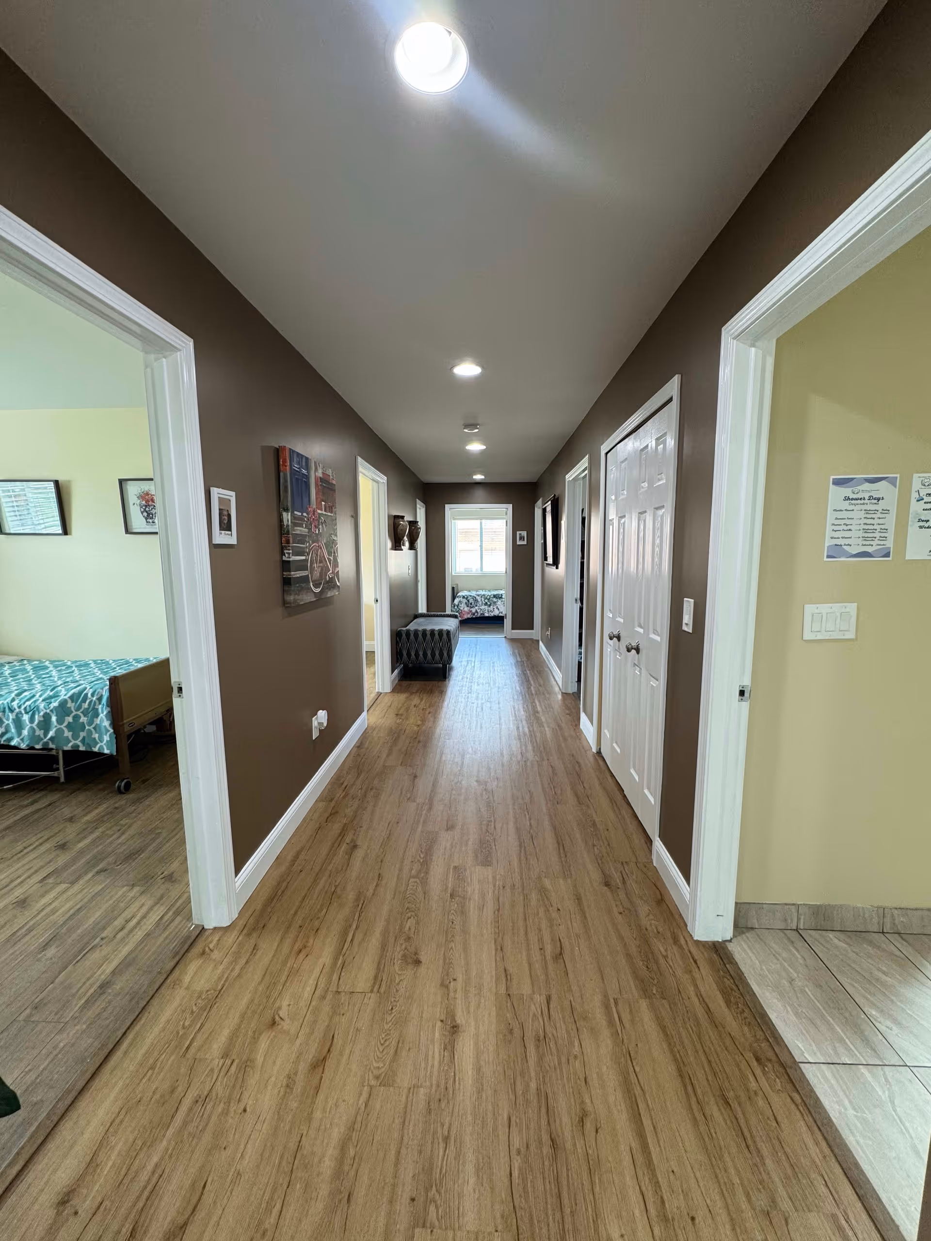 A long hallway with wood flooring and brown walls, featuring multiple doorways on both sides. On the left, a bedroom with a bed covered in a teal and white patterned blanket is visible. On the right, a room with tiled flooring and some posted signs on the wall can be seen. The hallway is lit by ceiling lights and has a bench near the end, with a window letting in natural light.