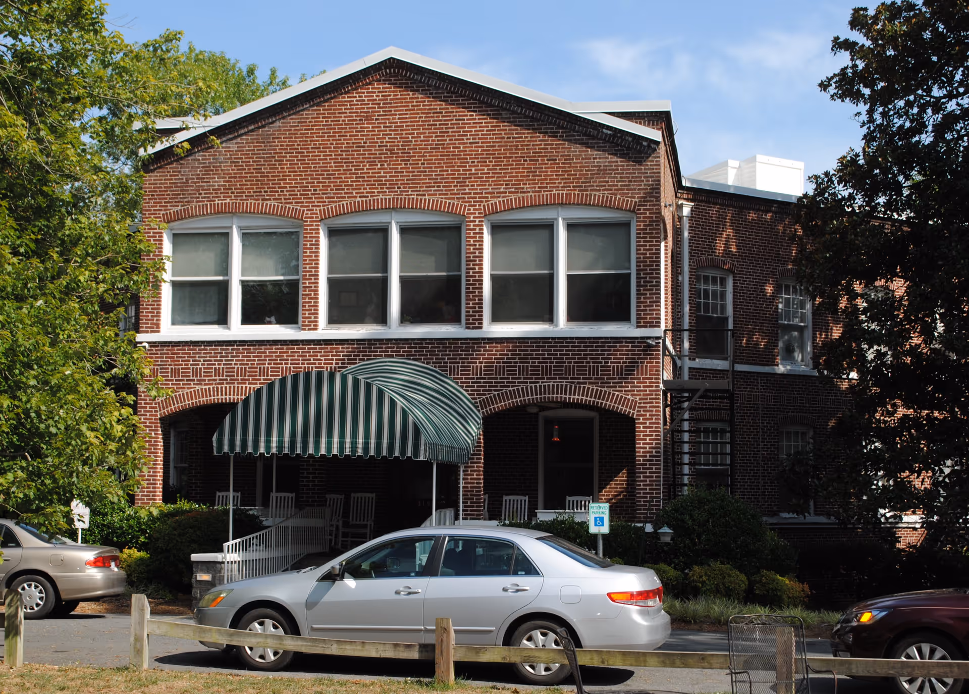 Front exterior view of a two-story brick building with white-framed windows and a green and white striped awning over the entrance. Several cars are parked in front, and there are trees and bushes around the building.