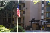 Exterior view of a multi-story senior living facility building with balconies, an American flag on a flagpole in front, and a covered entrance area surrounded by greenery.