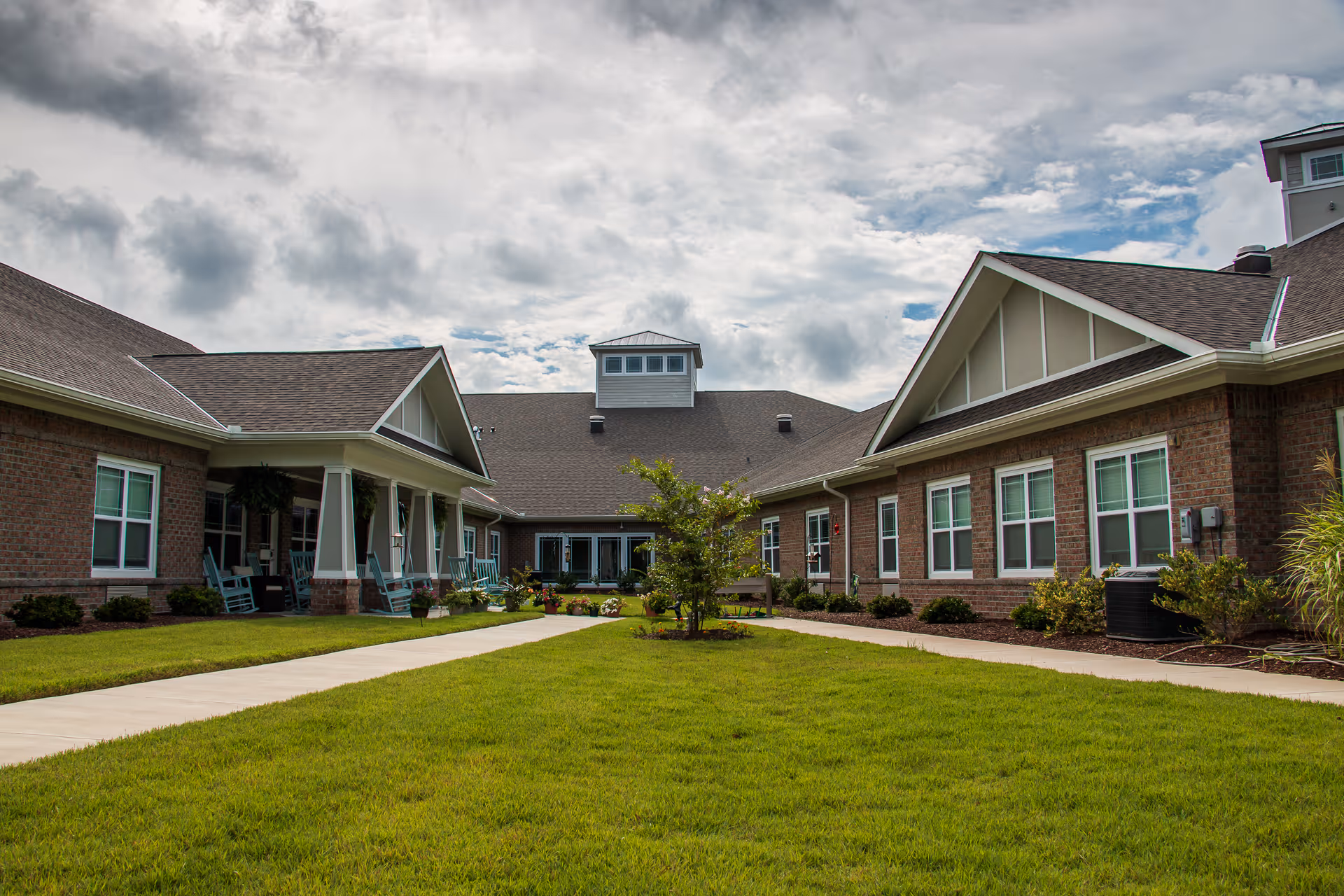 Brick buildings with porches and rocking chairs flank a green courtyard and walkway under a cloudy sky.