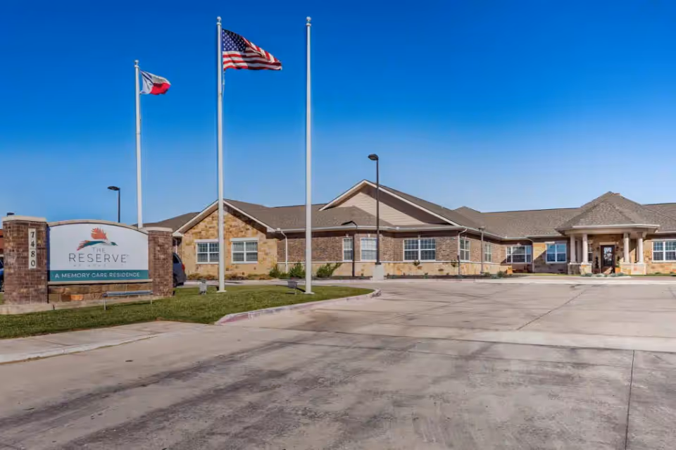 Exterior view of a single-story memory care residence building with a stone and brick facade under a clear blue sky. Three flagpoles display the Texas state flag, the American flag, and another flag. A sign near the entrance reads 'The Reserve at West Amarillo, A Memory Care Residence.' The driveway and parking area are visible in the foreground.