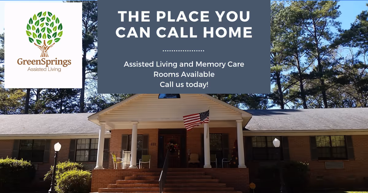 Front exterior view of Greensprings Assisted Living building with a porch, steps leading up to the entrance, an American flag, and trees in the background. The image includes a logo of a tree with green leaves and text promoting assisted living and memory care rooms available.