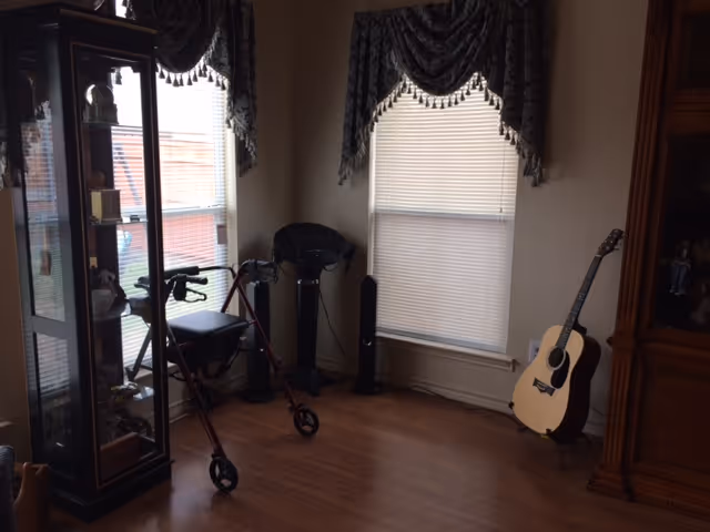 A corner of a room with wooden flooring, two windows with blinds and decorative valances, a black walker with a seat, a black standing fan, a wooden display cabinet with glass doors containing various items, and an acoustic guitar resting against the wall.