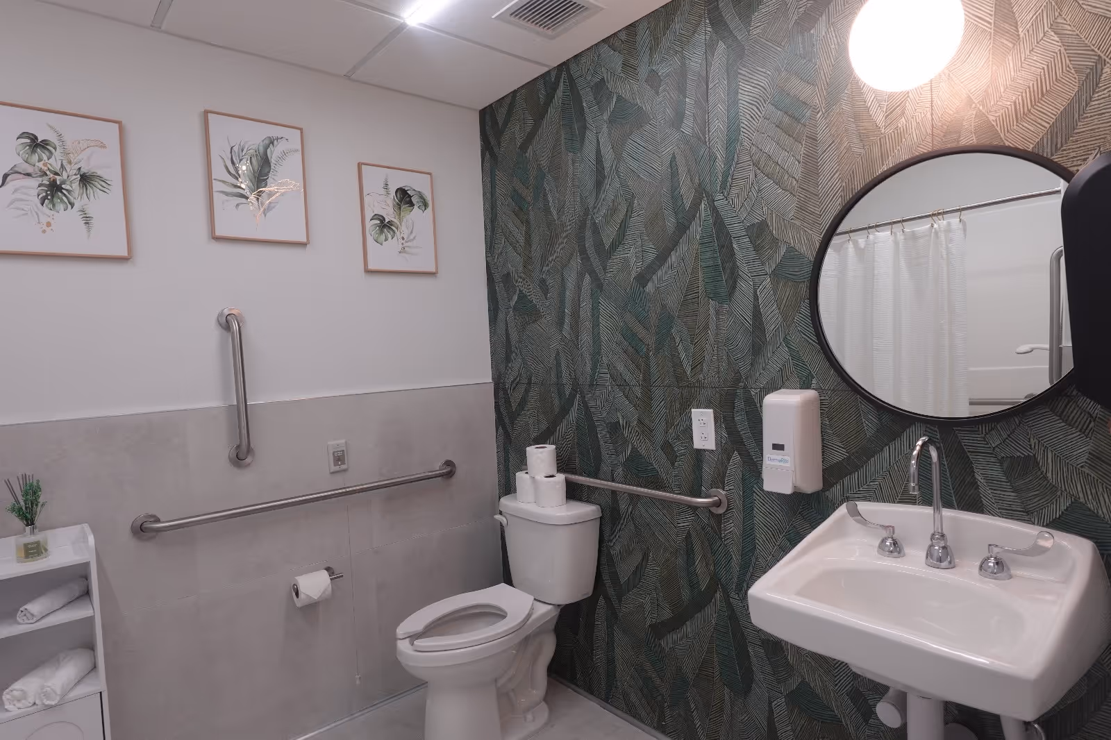 Accessible bathroom with a toilet, wall-mounted sink, round mirror, grab bars, and green leaf-patterned wallpaper.