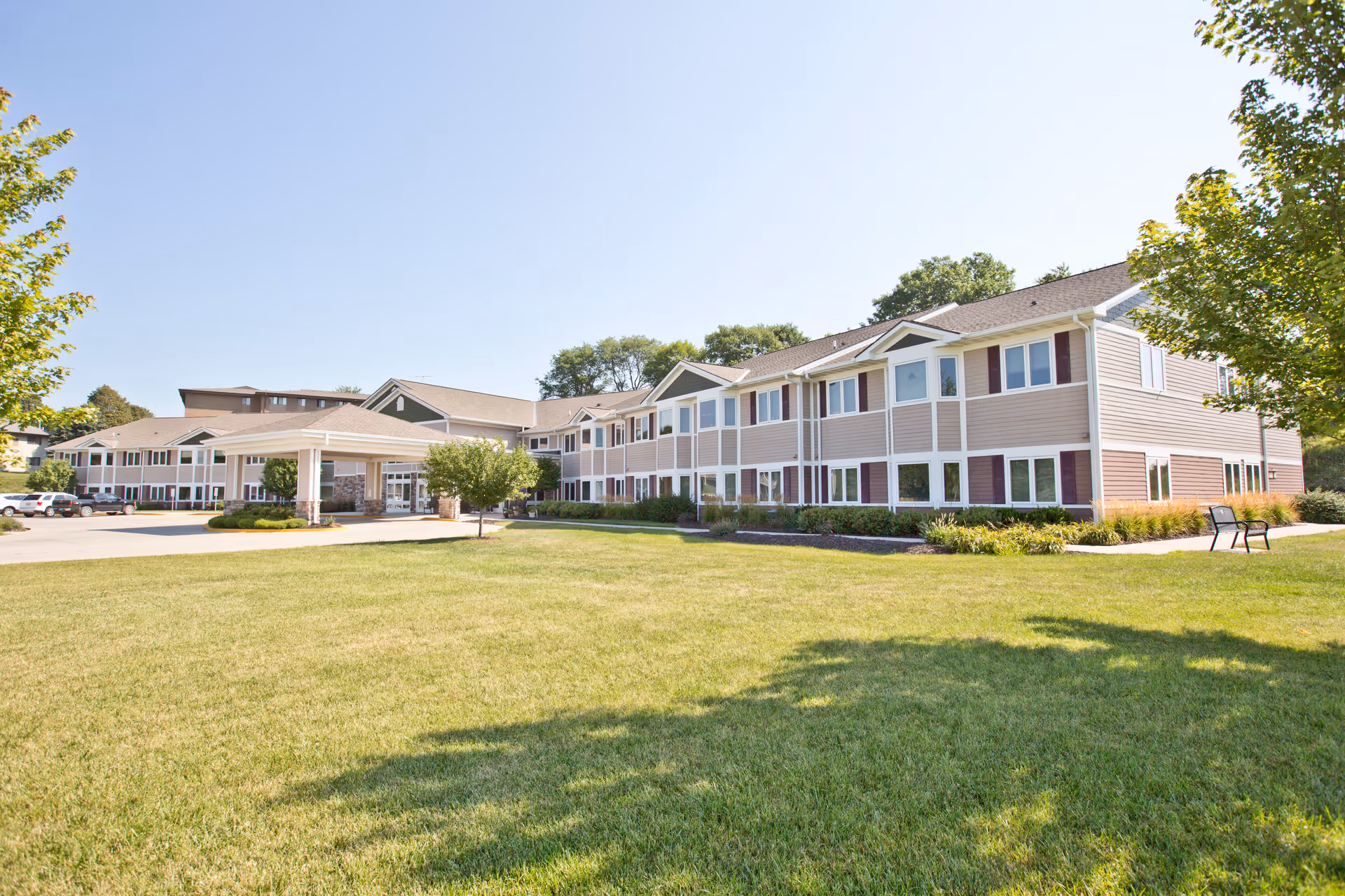 Exterior view of Silveridge Assisted Living, An Eventide Community, showing a large two-story building with beige siding and white trim, surrounded by a well-maintained lawn and trees under a clear blue sky.