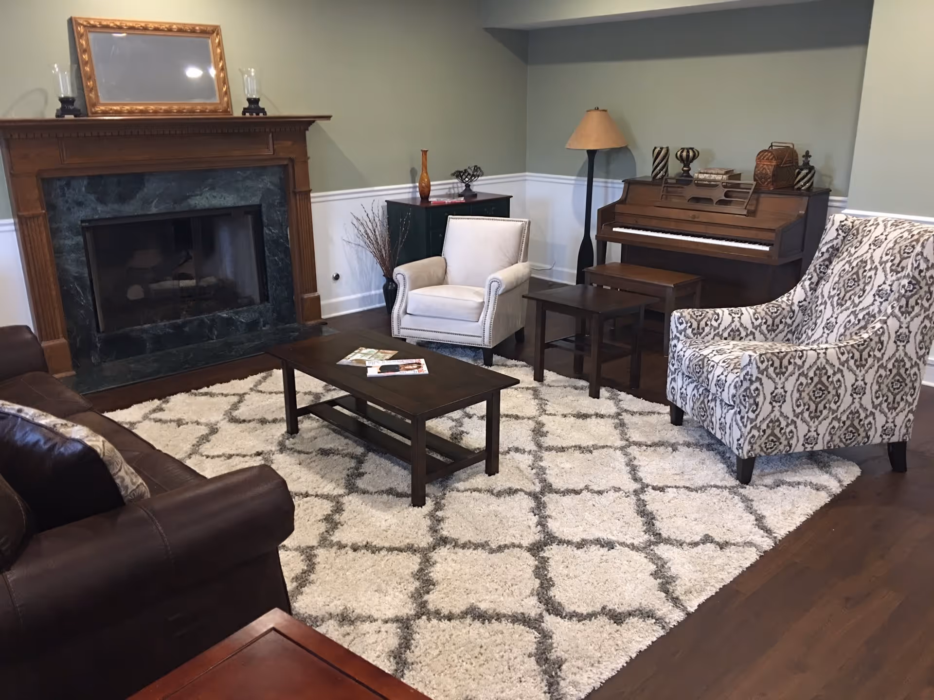 A cozy living room with a dark brown leather sofa, a white armchair, a patterned armchair, a wooden coffee table with magazines on it, a fireplace with a dark green marble surround and a wooden mantel, a wooden piano with decorative items on top, a floor lamp, and a light-colored rug with a geometric pattern on a wooden floor.