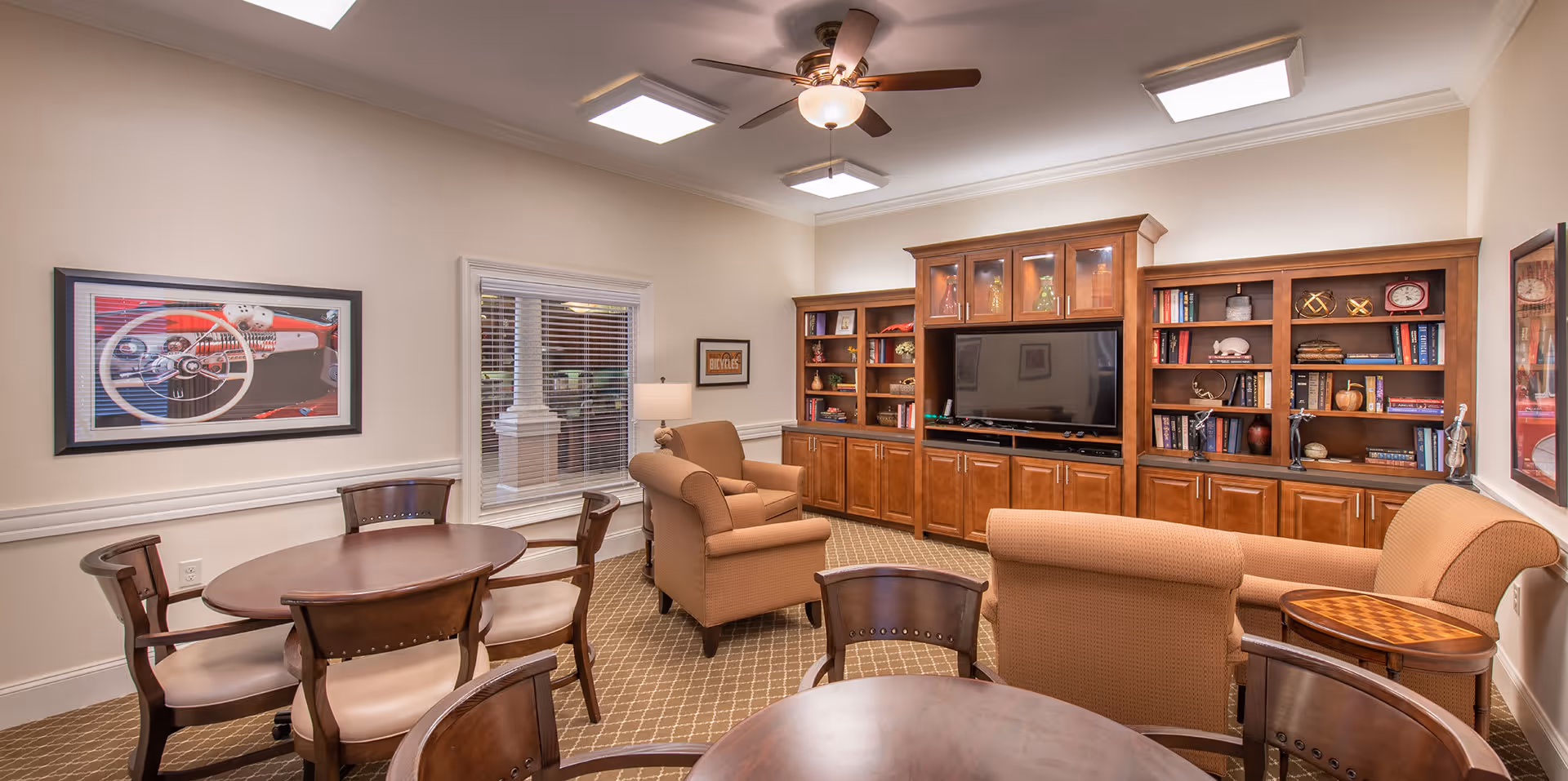 A cozy common living room with upholstered chairs and couches, round tables, a large TV set within wooden built-in bookshelves, and framed artwork on the walls.