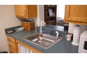 A kitchen countertop with a stainless steel sink, a faucet, wooden cabinets above and below the counter, a microwave on the right, a paper towel holder, a coffee mug, and a small basket on the left side of the counter.