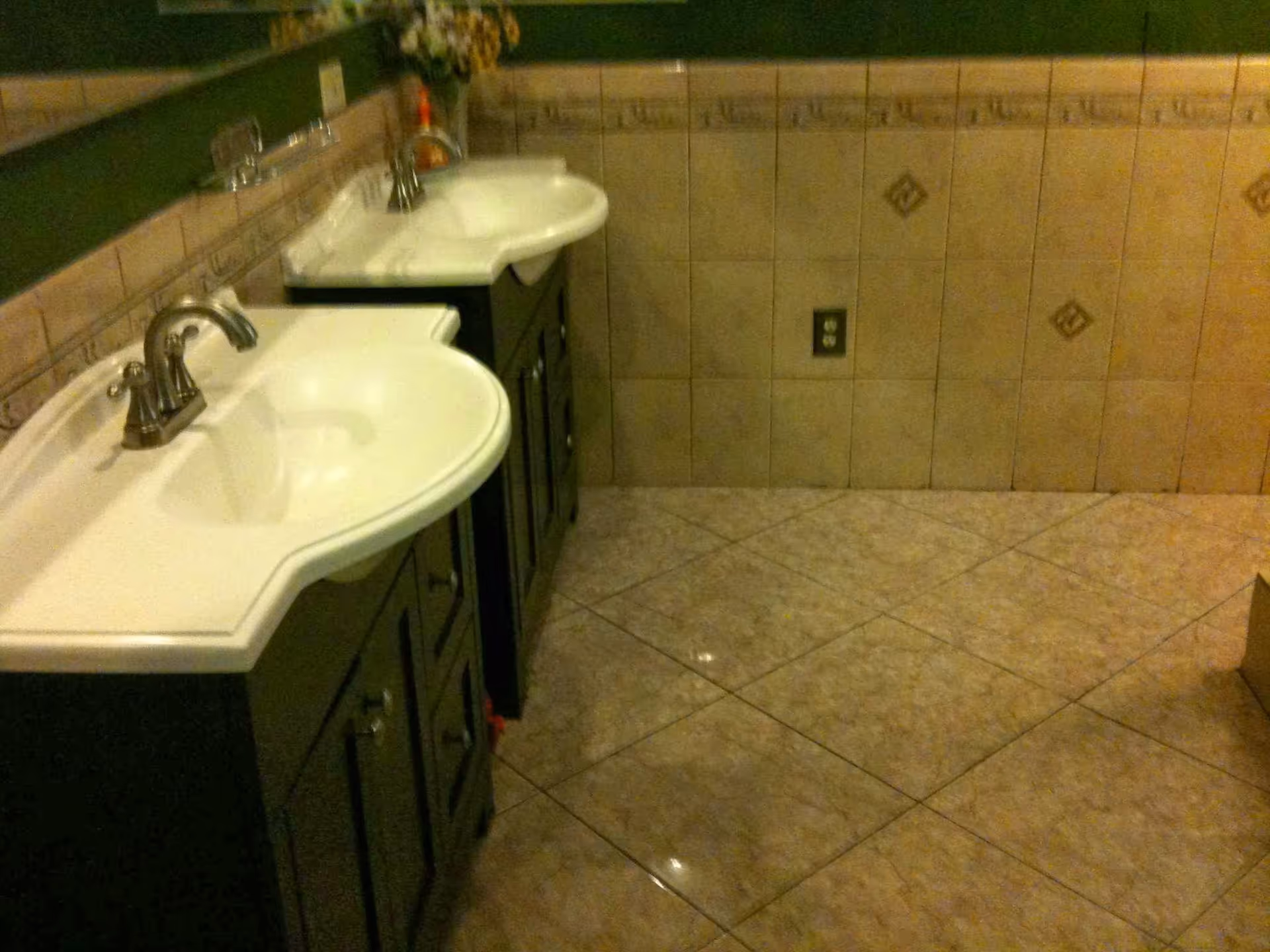 A bathroom interior featuring two white sinks with silver faucets mounted on dark wooden cabinets. The walls and floor are tiled with beige tiles, and there is a large mirror above the sinks. A vase with flowers is placed on the far sink.