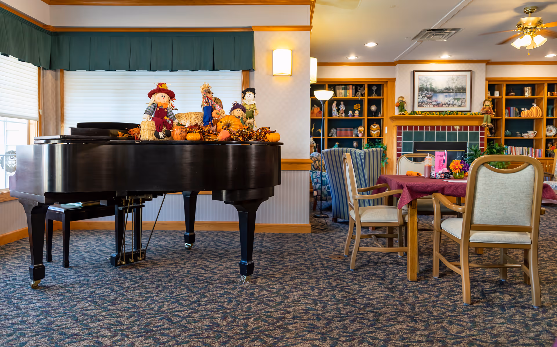 A cozy living room area in an assisted living facility featuring a black grand piano decorated with autumn-themed scarecrows and pumpkins. The room has carpeted flooring, a table with chairs covered with a maroon tablecloth, a fireplace with a green and red tile surround, built-in wooden bookshelves filled with books and decorative items, and a ceiling fan with lights.
