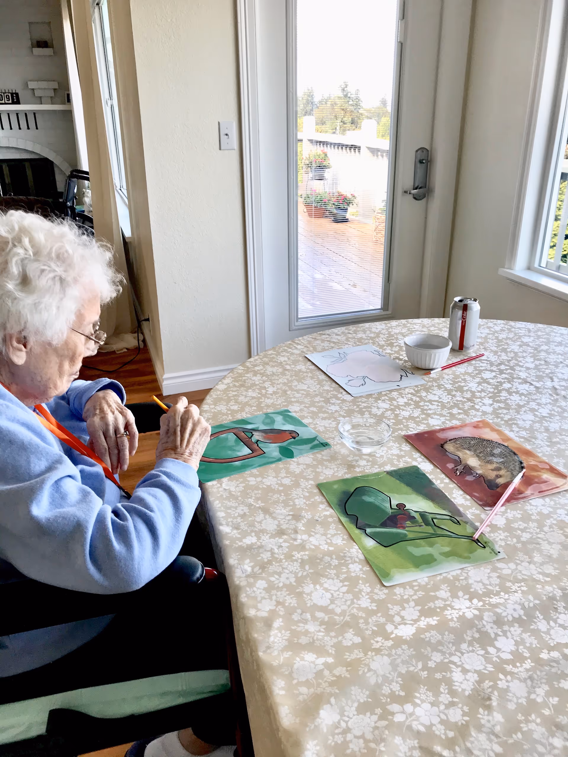 An elderly woman with white hair and glasses is seated at a round table covered with a floral tablecloth, engaged in coloring or drawing on a piece of paper. The table has several colorful drawings, a small bowl, and a can of Diet Coke. The room has light-colored walls, a door with a window leading outside, and a window letting in natural light.
